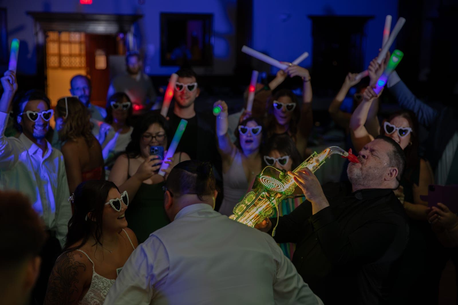 Glenn Frank playing a glowing gold LED saxophone on a packed dance floor with guests in heart-shaped sunglasses waving foam glow sticks, bride smiling in the foreground