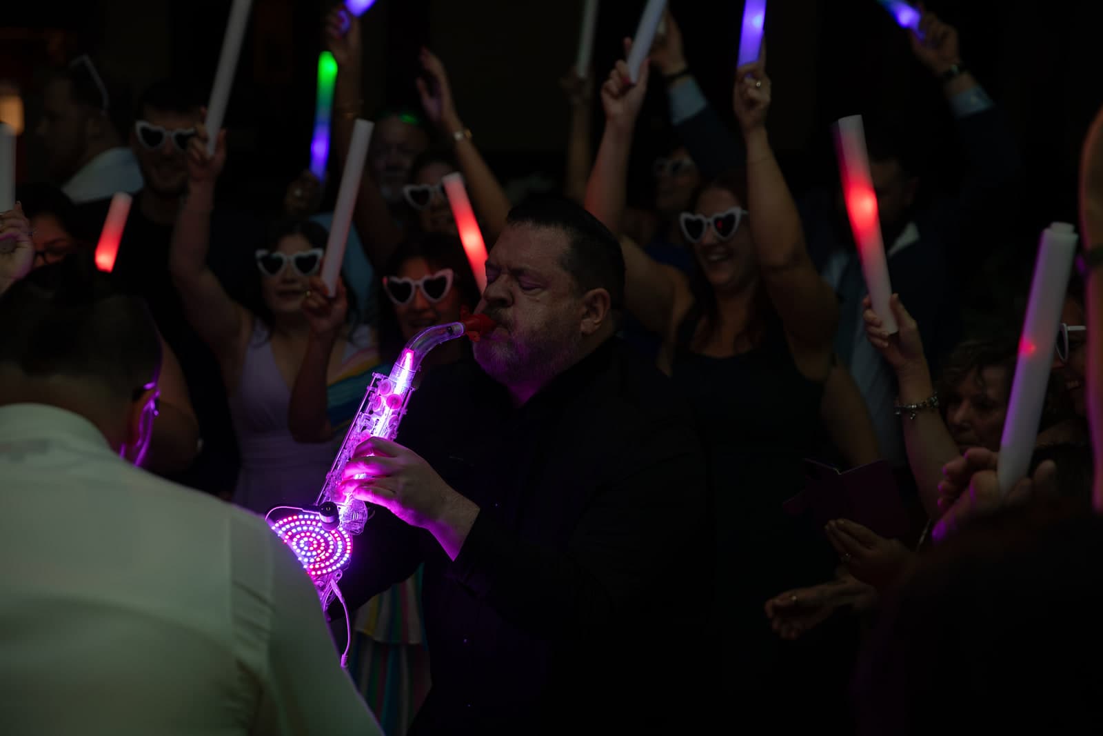 Glenn Frank performing on a pink and purple LED saxophone with guests in heart sunglasses and glow sticks behind him
